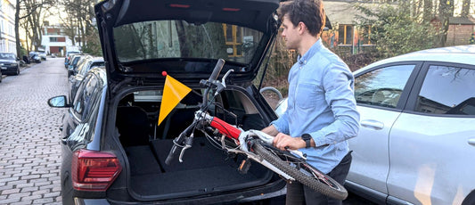 Mann mit Kinderfahrrad und Fahrradwimpel steht vor Kofferraum eines Autos